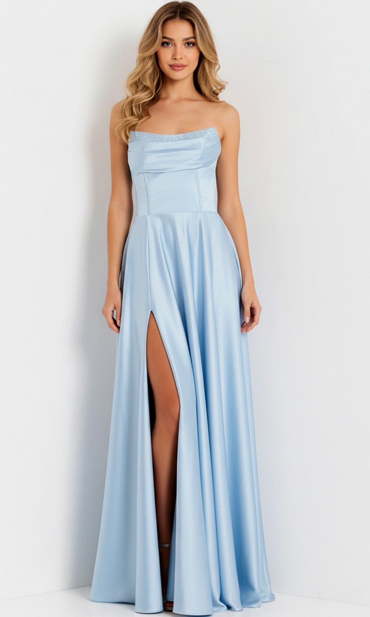 A woman stands against a white wall wearing the Jovani 46483 long prom dress, a light blue, sleeveless gown featuring a thigh-high slit, fitted bodice, and delicate beaded trim.